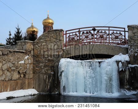 The Frozen Waterfall Of The Holy Water Under The Footbridge. Church. Golden Domes.