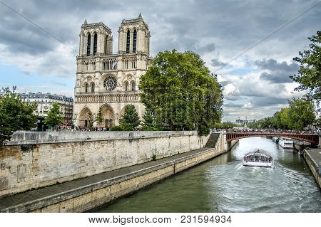 View Of Notre-dame Cathedral On City Sena River Bank With Cloudy Sky And Green Trees Among