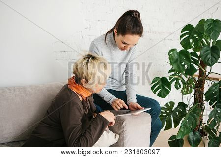 A Young Girl Volunteer Or Granddaughter Explains To An Elderly Woman How To Use A Tablet Or Shows So