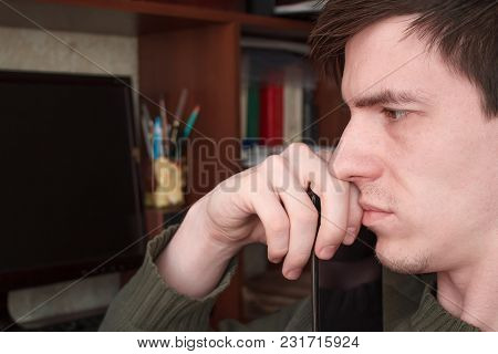 A Thoughtful Guy With A Smartphone At His Mouth, Sits At His Desk, On A Black Monitor Background