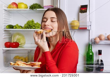 Photo Of Hungry Young Cute Woman Eats With Appetite Tasty Sausage Sandwhich, Comes After Work, Stand
