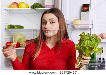 Hesitant Young Female Model Holds Lettuce And Sandwhich In Hands, Doesn`t Know What To Choose, Can`t