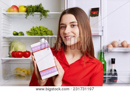 Photo Of Good Looking Young Female Dressed In Red Blouse, Holds Pack Of Milk, Stands Near Fridge Ful