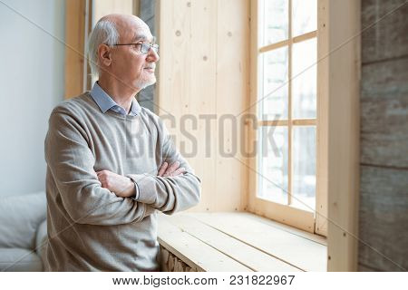 Pensive Mood. Thoughtful Attractive Senior Man Gazing Through Window While Crossing Hands And Stayin