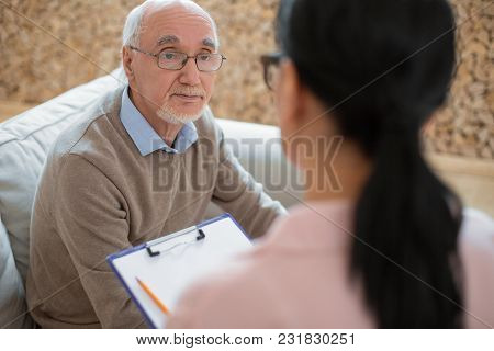 First Meeting. Pleasant Experienced Senior Man Posing On Sofa While Staring At Caregiver And Communi
