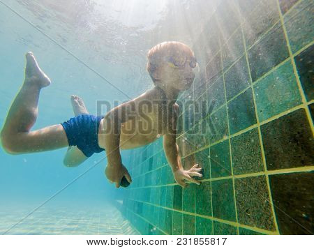 A Child Boy Is Swimming Underwater In A Pool, Smiling And Holding Breath, With Swimming Glasses.