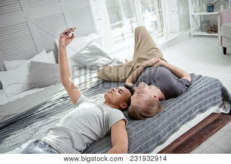 Smile For Photo. Nice Appealing Gay Couple Lying On Bed While Shooting Selfie And Resting