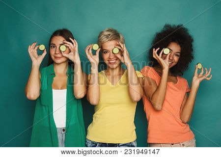 Three Beautiful Girls In Colourful Clothes Having Fun, Covering Eyes With Cucumber Pieces On Green B