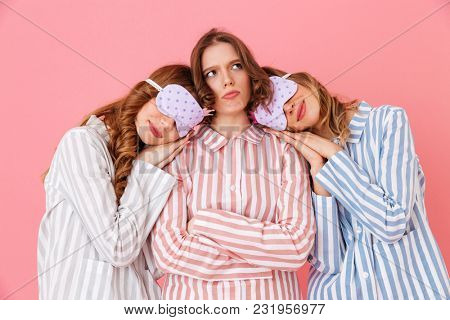 Three friendly young girls 20s wearing colorful striped pyjamas and sleeping masks sleeping while playing around during girlish sleepover isolated over pink background