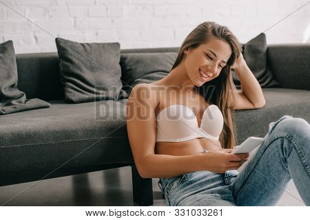 Smiling Woman In White Bra And Jeans Using Smartphone While Sitting On Floor Near Sofa