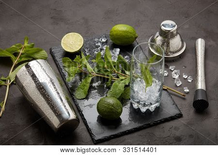 Set of bar tools: cocktail shaker, muddler and glass with ice, lime and mint  on dark stone  table. Top view. Bartender's tool. Mojito cocktail making.