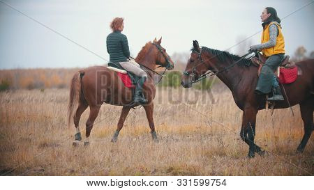 Two Women In Jackets Riding Horses On The Autumn Field