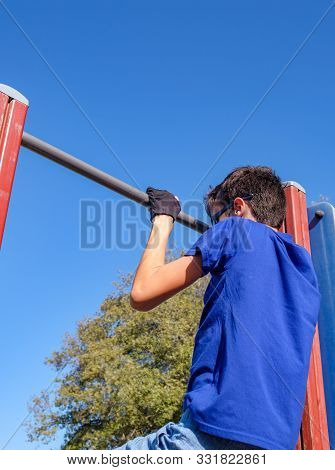 A Boy Pulls Himself Up On The Horizontal Bar On Blue Sky Background. Teenager Wears Suede Leather Gr
