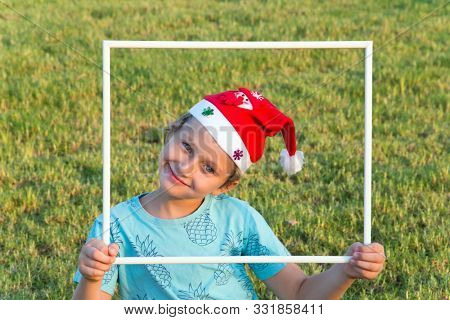 Charming boy in a clownish cap of Santa Claus smiles through a white frame. Concept portrait and advertising photo. The background is green lawn