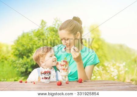 Mother And Her Son Child Boy Are Eating Laered Dessert In Sunny Day In Green Garden