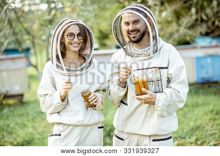 Portrait Of A Man And Woman Beekepers In Protective Uniform Standing Together With Honey In The Jar,