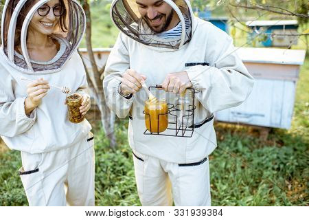 Two Beekepers In Protective Uniform Standing Together With Honey In The Jar, Tasting Fresh Product O