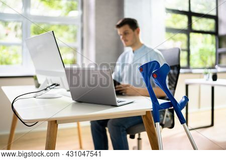 Handicapped Injured Employee With Crutches At Workplace Desk