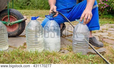 Man In A Blue Jumpsuit Pours Drinking Water From A Hose Into Large Bottles. Eco-friendly Drinking Wa