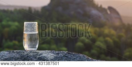 A Glass Of Pure Transparent Mineral Drinking Water Against The Background Of A Sunset In The Mountai