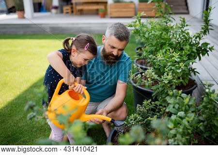 Father And Small Daughter With Watering Can Outdoors In Tha Backyard, Looking At Camera.
