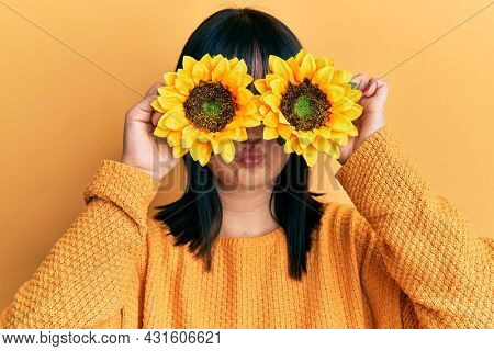 Young hispanic woman holding sunflowers over eyes puffing cheeks with funny face. mouth inflated with air, catching air. 