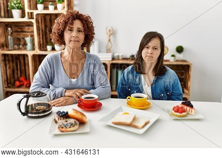 Family of mother and down syndrome daughter sitting at home eating breakfast relaxed with serious expression on face. simple and natural looking at the camera. 
