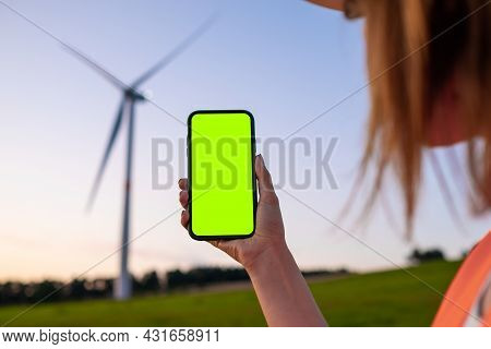 Woman Engineer Holding A Smartphone With Chromo Key On The Background Of Wind Turbine At Sunset.