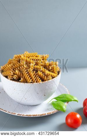 Raw Fusilli Pasta In A White Bowl With Cherry Tomatoes And Basil On A Gray Background. Whole Grain P