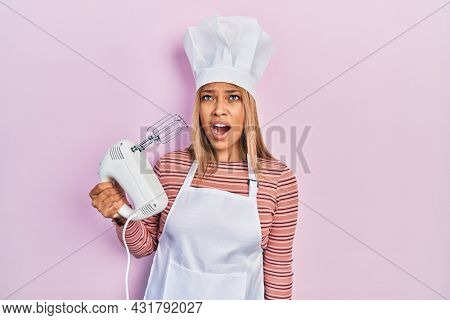 Beautiful hispanic woman holding pastry blender electric mixer angry and mad screaming frustrated and furious, shouting with anger. rage and aggressive concept. 