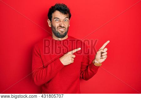 Handsome man with beard wearing casual red sweater pointing aside worried and nervous with both hands, concerned and surprised expression 