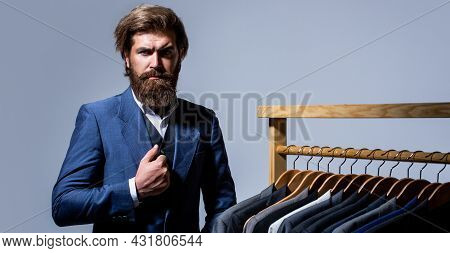 Handsome Bearded Fashion Man In Classical Costume Suit. Male Suits Hanging In A Row. Men Clothing, B