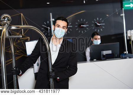 Bellboy In Medical Mask Pushing Luggage Cart Near Blurred Administrator In Hotel Lobby