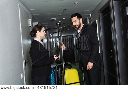Smiling Receptionist Pointing With Hand Near Bellboy Near Luggage Cart