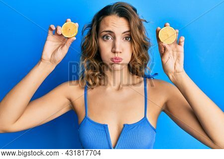 Young caucasian woman wearing swimsuit holding slices of lemon over eyes puffing cheeks with funny face. mouth inflated with air, catching air. 