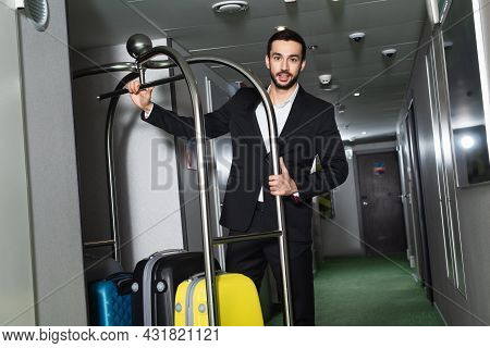 Bearded Bellboy Near Metallic Bell Cart With Luggage In Hall Of Hotel