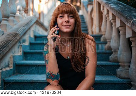 Portrait Of Young Smiling Woman With Tattoos Is Sitting On The Stome Stairs And Talking On The Phone