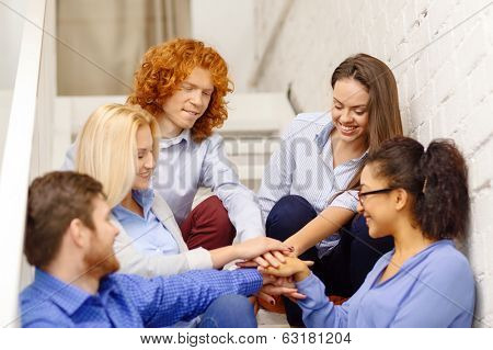 business, office, gesture and startup concept - smiling creative team with hands on top of each other sitting on staircase