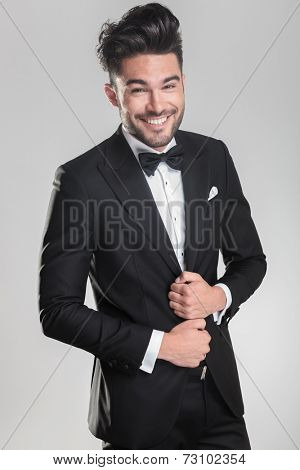 Happy young man wearing a tuxedo smiling for the camera while ajusting his jacket. on a grey background.