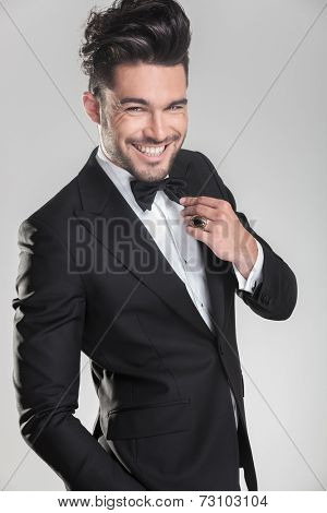 Elegant young man in tuxedo ajusting his bow tie while smiling for the camera. On grey background.