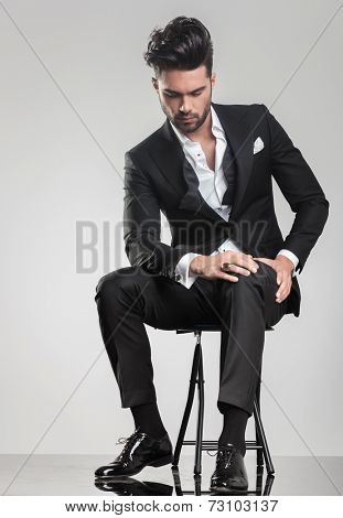 Elegant young man in tuxedo stitting on a stool while looking down, holding one hand on hei knee.