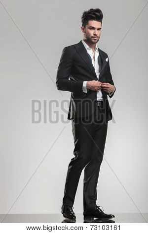 Full body image of an handsome young man looking away from the camera while closing his tuxedo. 