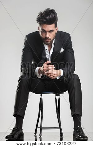 Elegant fashion man in tuxedo looking at the camera while sitting on a stool, holding his hands.