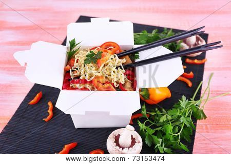 Chinese noodles and sticks in takeaway box on black mat on pink background