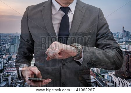 Double exposed of Businessman checking time on his wrist watch and holding mobile phone with cityscape.