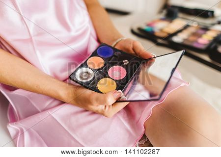 Young beautiful woman holds in two hand and a palette with paint and shadows with mirror for makeup