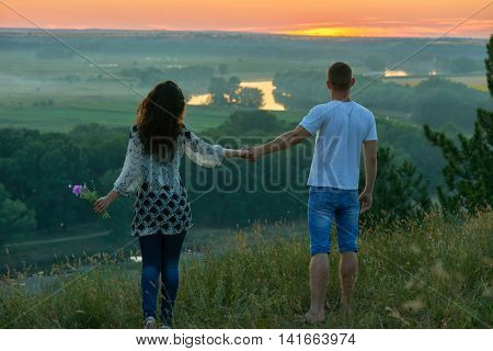 romantic couple walk on hill at sunset on country outdoor, girl with bouquet of wild flower, beautiful landscape and bright yellow sky, love tenderness concept, young adult people