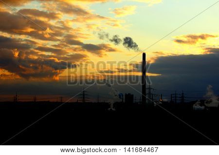 A silhouette of a power plant with a high smoking chimney at a sunset