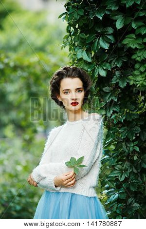 Portrait of a young slim beautiful girl with retro hair wearing a white blouse blue skirt posing against a background of vineyard