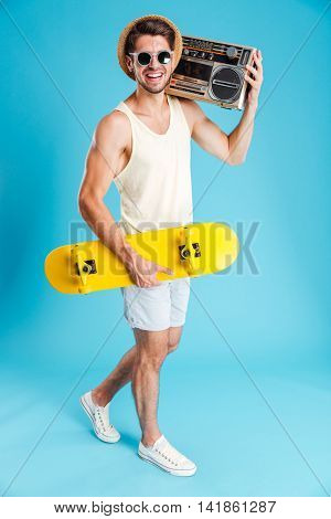 Smiling young man with yellow skateboard walking and holding old boombox over blue background